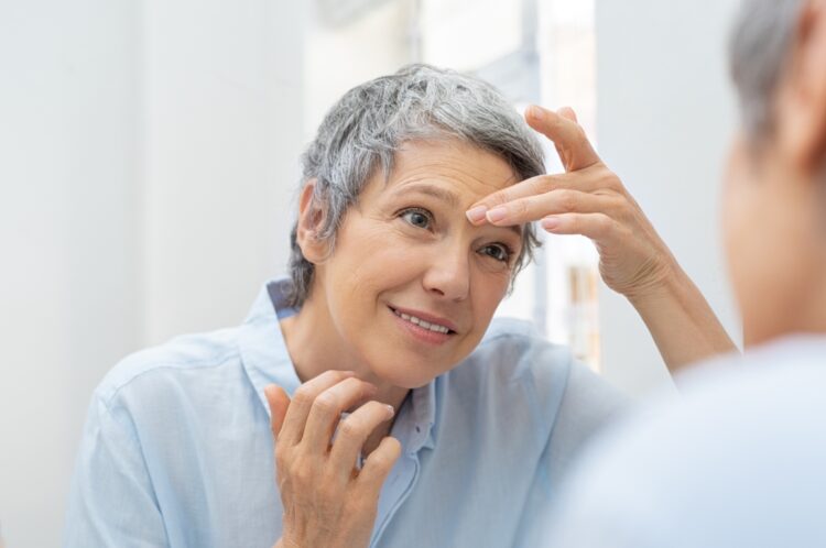 Mature woman looking her face and wrinkles in the bathroom mirror. Senior woman applying cosmetic lotion on skin between eyebrows while looking at mirror. Beautiful lady looking her face in the early morning.